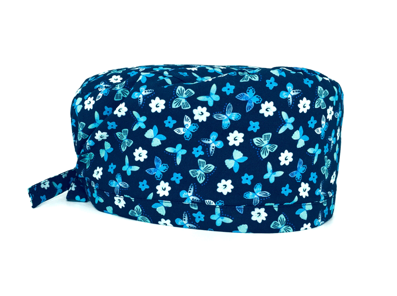 La base blu di questa cuffietta chirurgica esalta le sfumature di azzurro, turchese e verde acqua che colorano le ali di piccole farfalle e i petali di delicati fiorellini. 
