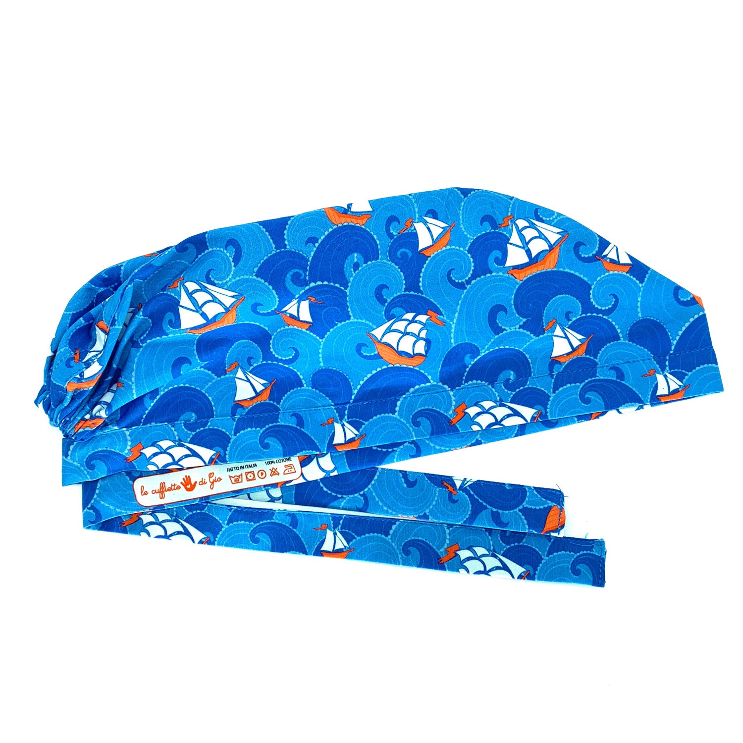 Cuffietta chirurgica con onde stilizzate alternate azzurro intenso e blu medio, fra le onde emergono piccole imbarcazioni e grandi velieri che danno un tocco di bianco e rosso. Tema marino stilizzato con colori classici blu rosso e bianco