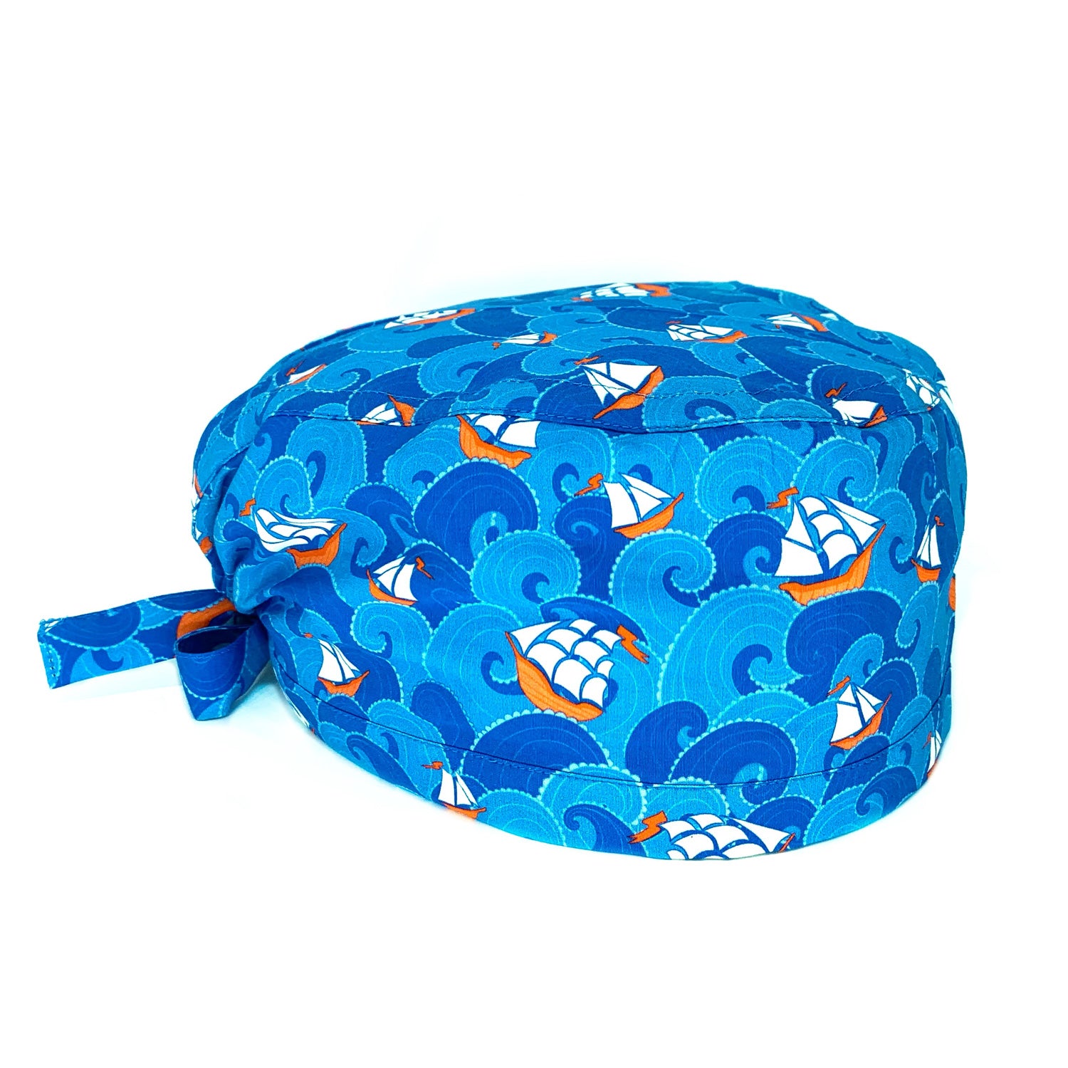 Cuffietta chirurgica con onde stilizzate alternate azzurro intenso e blu medio, fra le onde emergono piccole imbarcazioni e grandi velieri che danno un tocco di bianco e rosso. Tema marino stilizzato con colori classici blu rosso e bianco